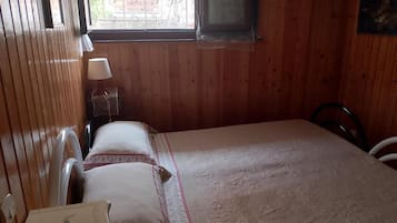 Una habitación con paredes de madera, una cama con cubrecama floral, una mesita de noche con lámpara y una ventana con vista a edificios.