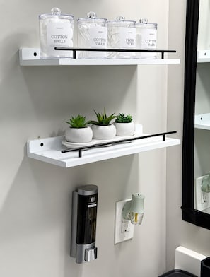 A bathroom with a white shelf holding cotton balls, swabs, floss picks, and cotton pads. Below, there are potted plants and a hand dryer.