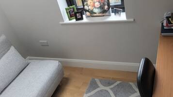 A room with a grey sofa, a wooden floor, a small rug, a black chair, and a shelf with framed photos and books.
