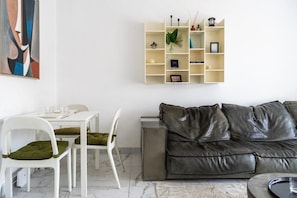 Ein modernes Wohnzimmer mit einem weißen Esstisch, zwei weißen Stühlen, einem grauen Ledersofa und einem weißen Bücherregal.