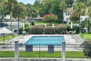 Ein Swimmingpool mit schwarzem Zaun, umgeben von Liegestühlen und Sonnenschirmen, mit Palmen und einem Gebäude im Hintergrund.