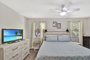 Une chambre à coucher avec un lit, une télévision, un ventilateur de plafond et une fenêtre avec des stores.