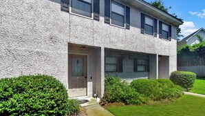 A two-story residential building with a textured facade, a front door with a glass panel, and a small porch area. - Great condo 1/2 mile to the pier village and 3/4 mile to the beach (Saint Simons Island)