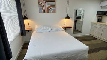 A neatly made bed in a hotel room with a bedside table, a wall-mounted mirror, and a small shelf with a lamp.