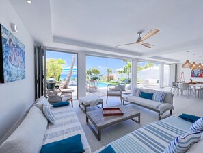 A modern living room with a pool view, large windows, and a ceiling fan. - Luxury villa · St-François · Sea view & beach access (Saint-François)