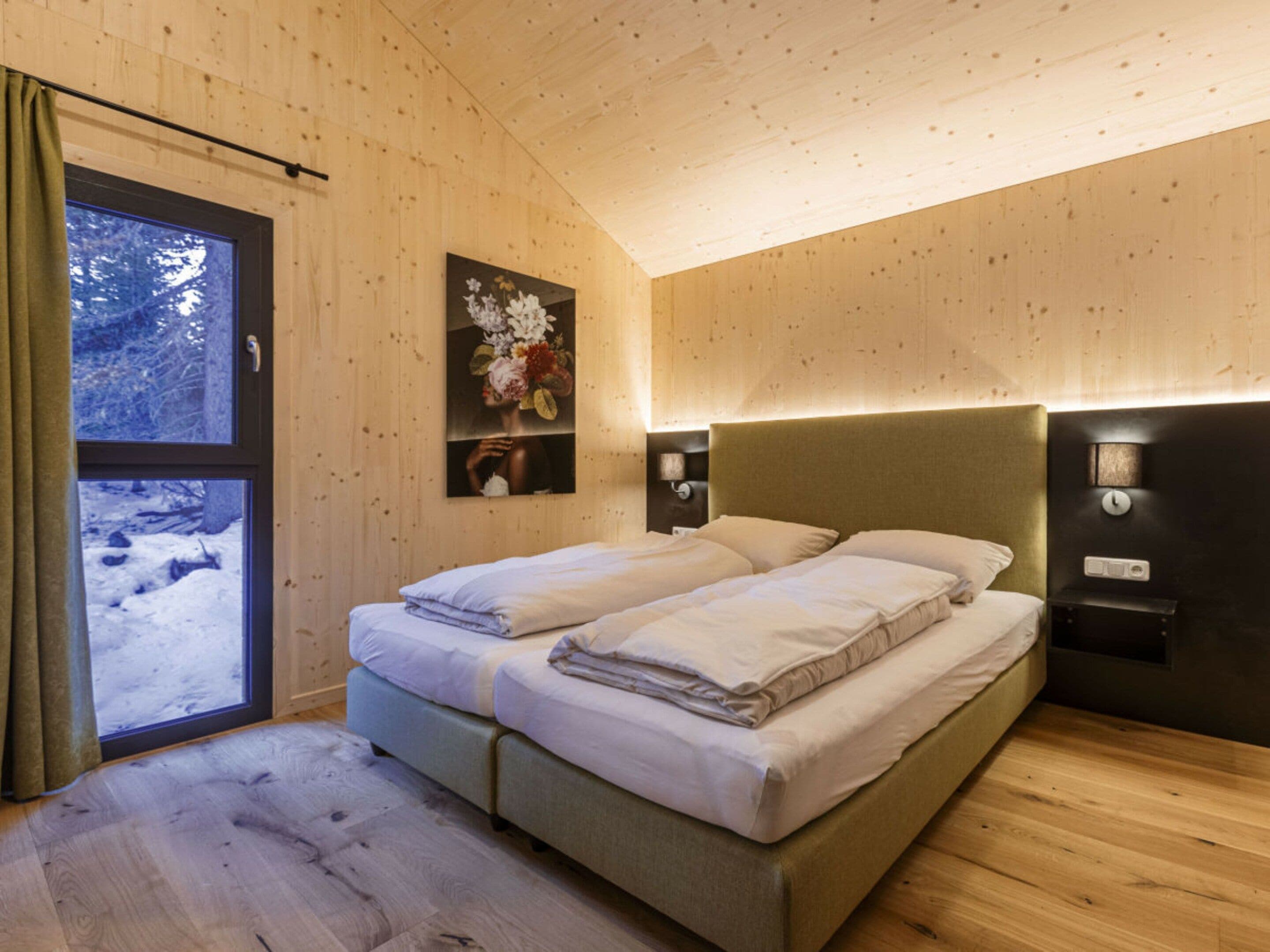 A room with wooden walls and a bed with two pillows. There is a window with curtains and a painting on the wall.
