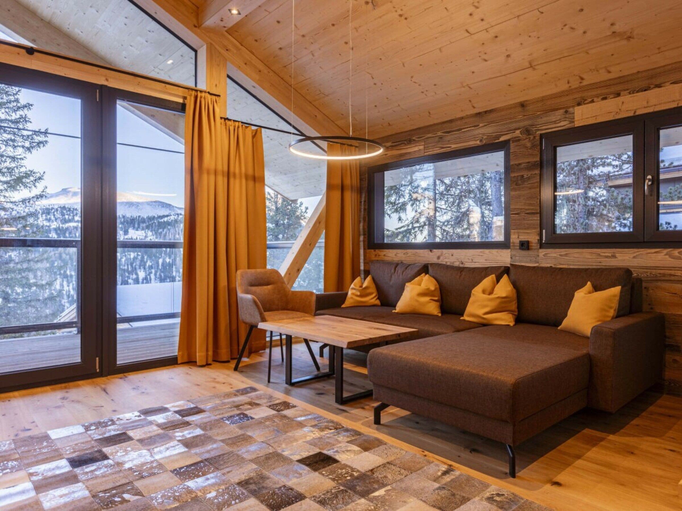 Een gezellig chaletinterieur met een houten vloer, een bruine bank met gele kussens, een houten tafel en grote ramen met uitzicht op met sneeuw bedekte bergen.