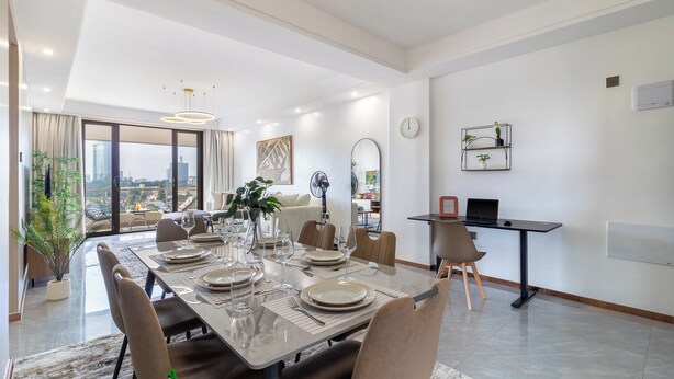 A modern dining area with a large table set for six, a city view, and a kitchen area with a laptop and a clock. - Luxury 4BR All-ensuite Heated Pool Riverside, Nairobi (Nairobi)