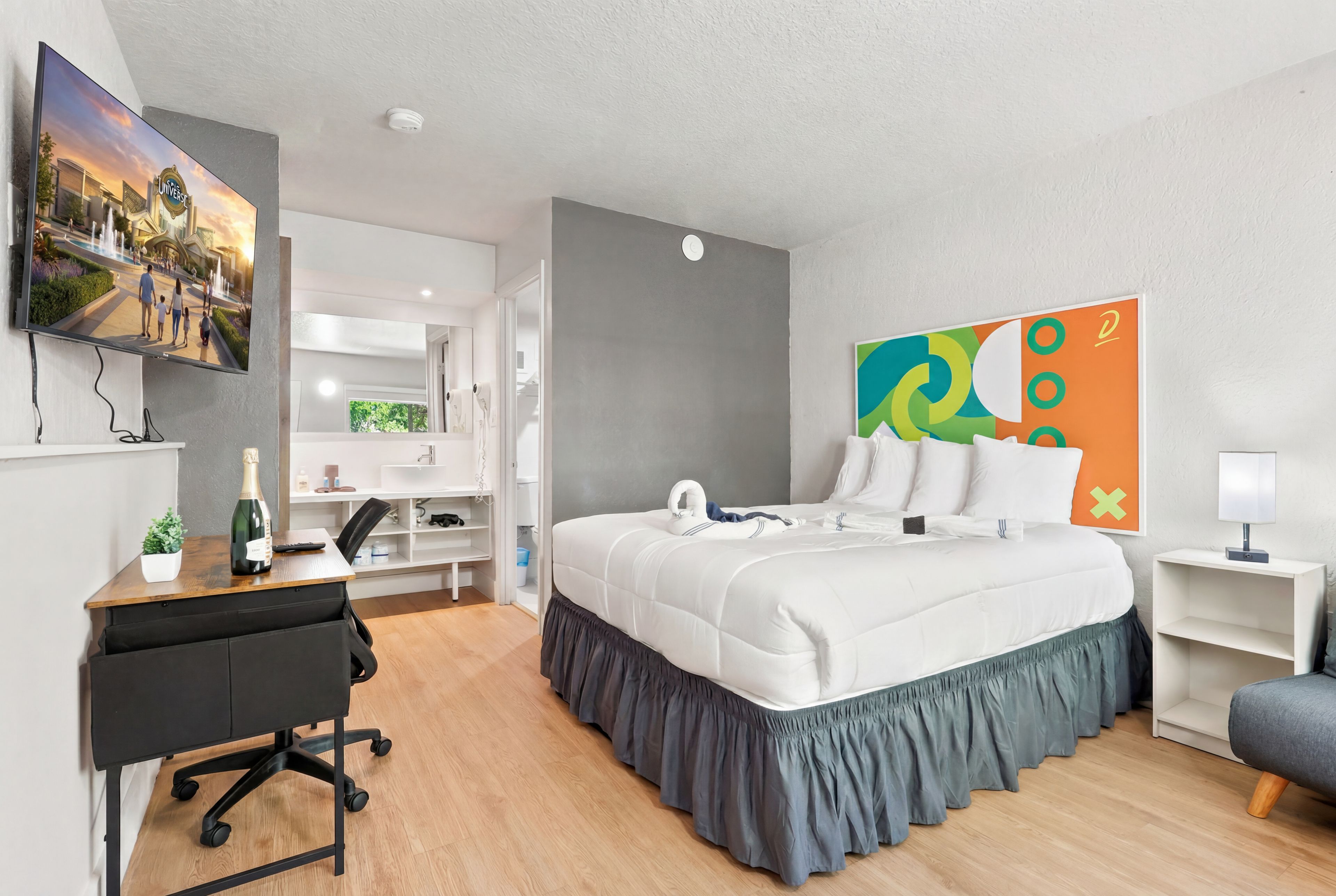 A hotel room with a bed, desk, chair, TV, and artwork.