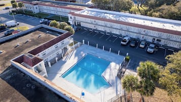An aerial view of a hotel with a swimming pool, parking area, and adjacent buildings.