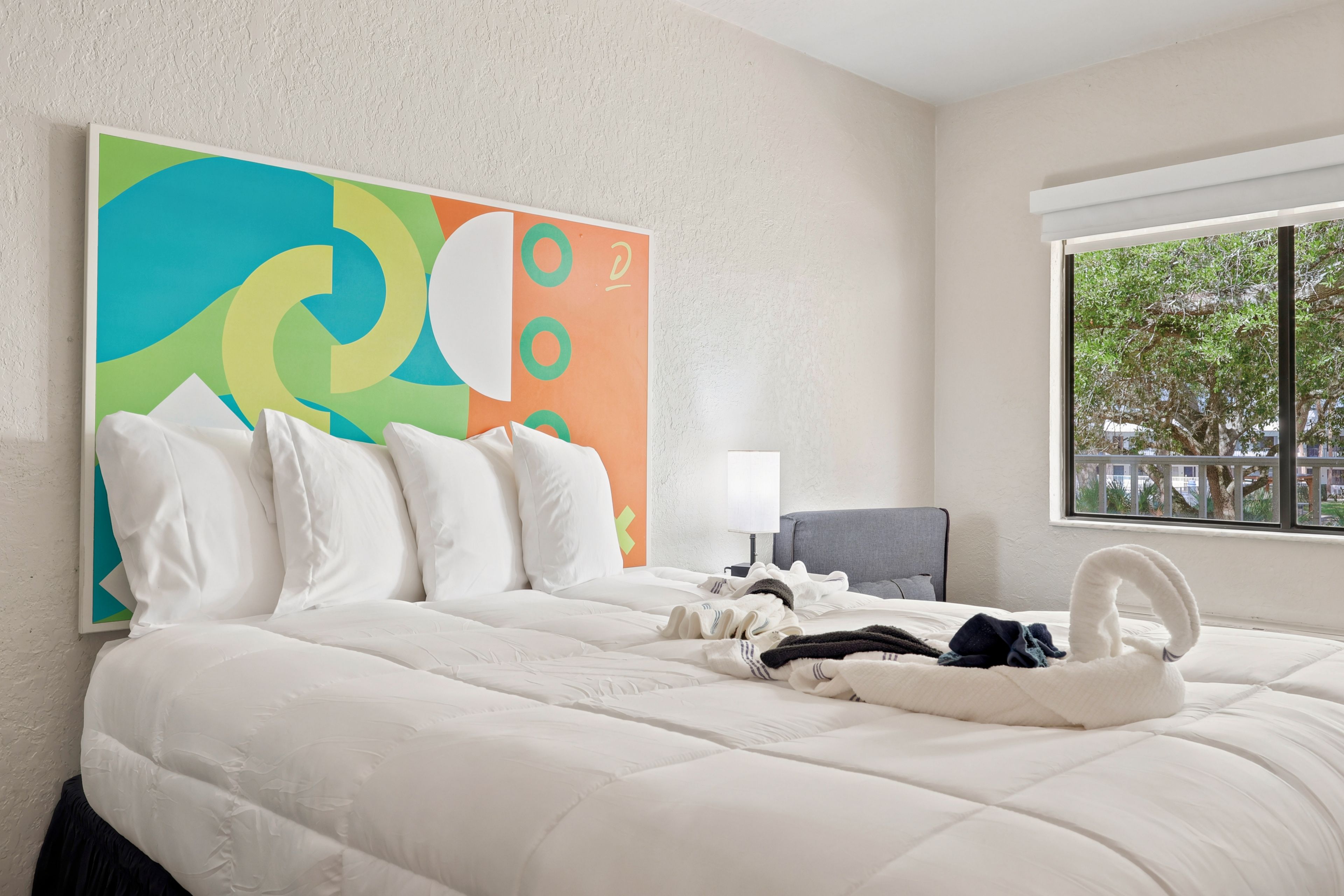 Une chambre d’hôtel avec un grand lit, un tableau abstrait aux couleurs vives, une fenêtre donnant sur un beau paysage et un fauteuil.