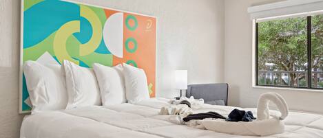 Une chambre d’hôtel avec un grand lit, un tableau abstrait aux couleurs vives, une fenêtre donnant sur un beau paysage et un fauteuil.