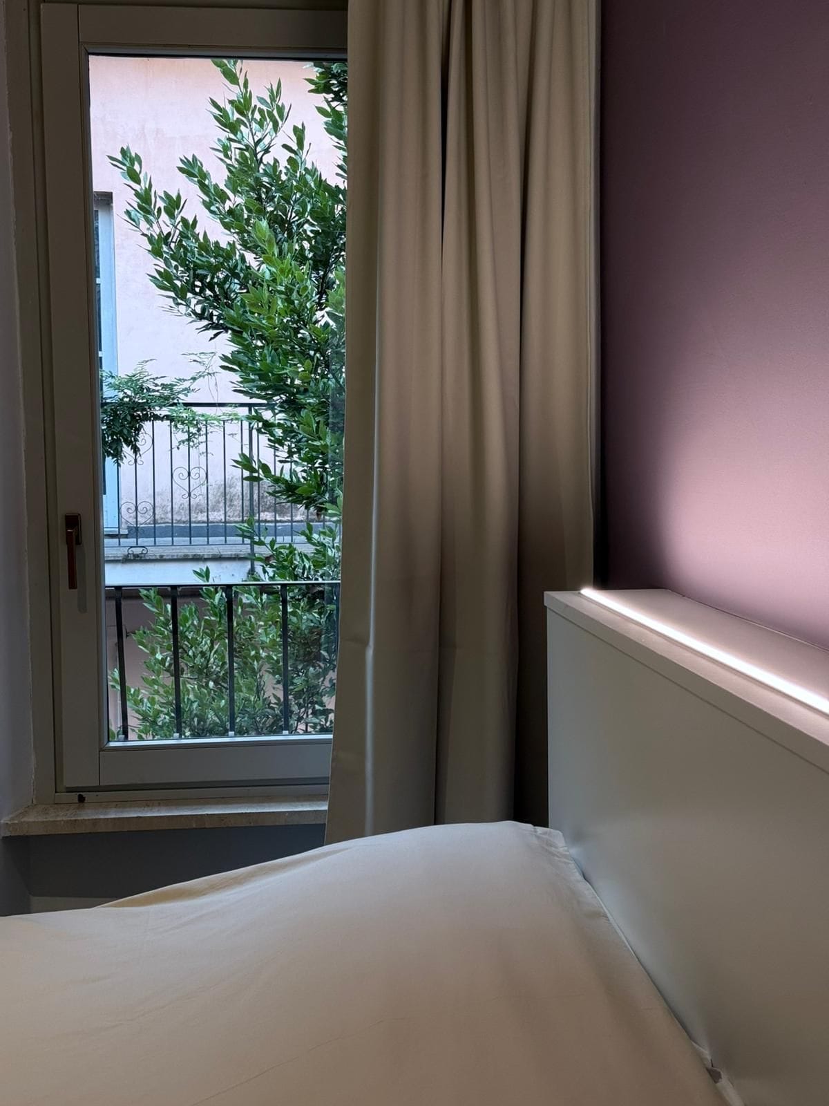 Une chambre avec un lit, une fenêtre avec des rideaux et une vue sur un balcon agrémenté de verdure et d’une clôture.