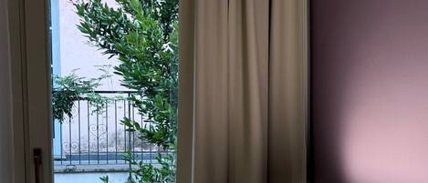 A room with a bed, a window with curtains, and a view of a balcony with greenery and a fence.