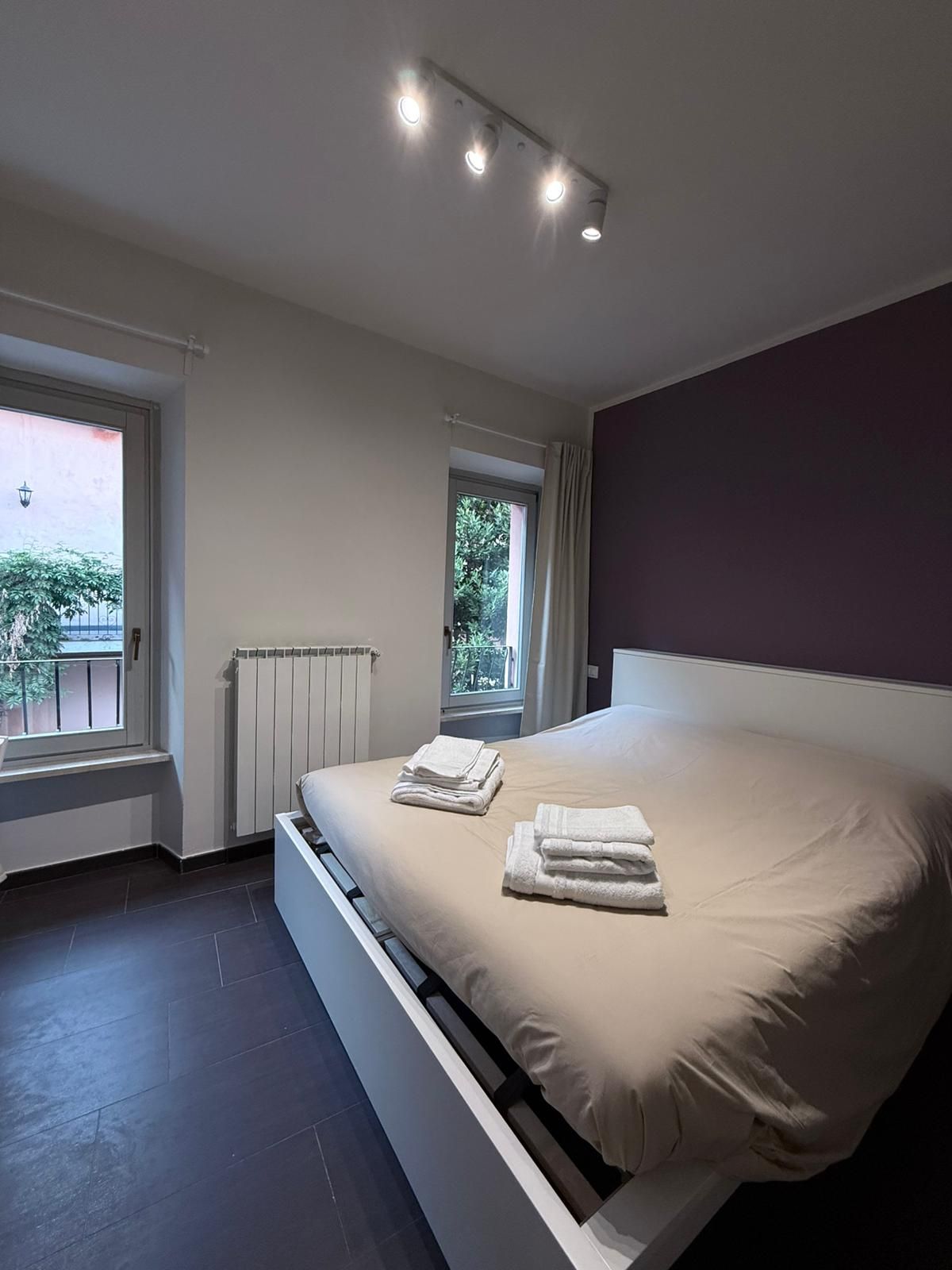 Un dormitorio moderno con una cama grande, dos ventanas, un radiador y una lámpara en el techo.