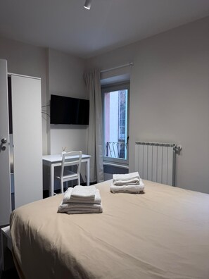A hotel room with a bed, a desk with a chair, a television mounted on the wall, and a window with a view of the outside. - Ametista | Urban Suite (Valenza)