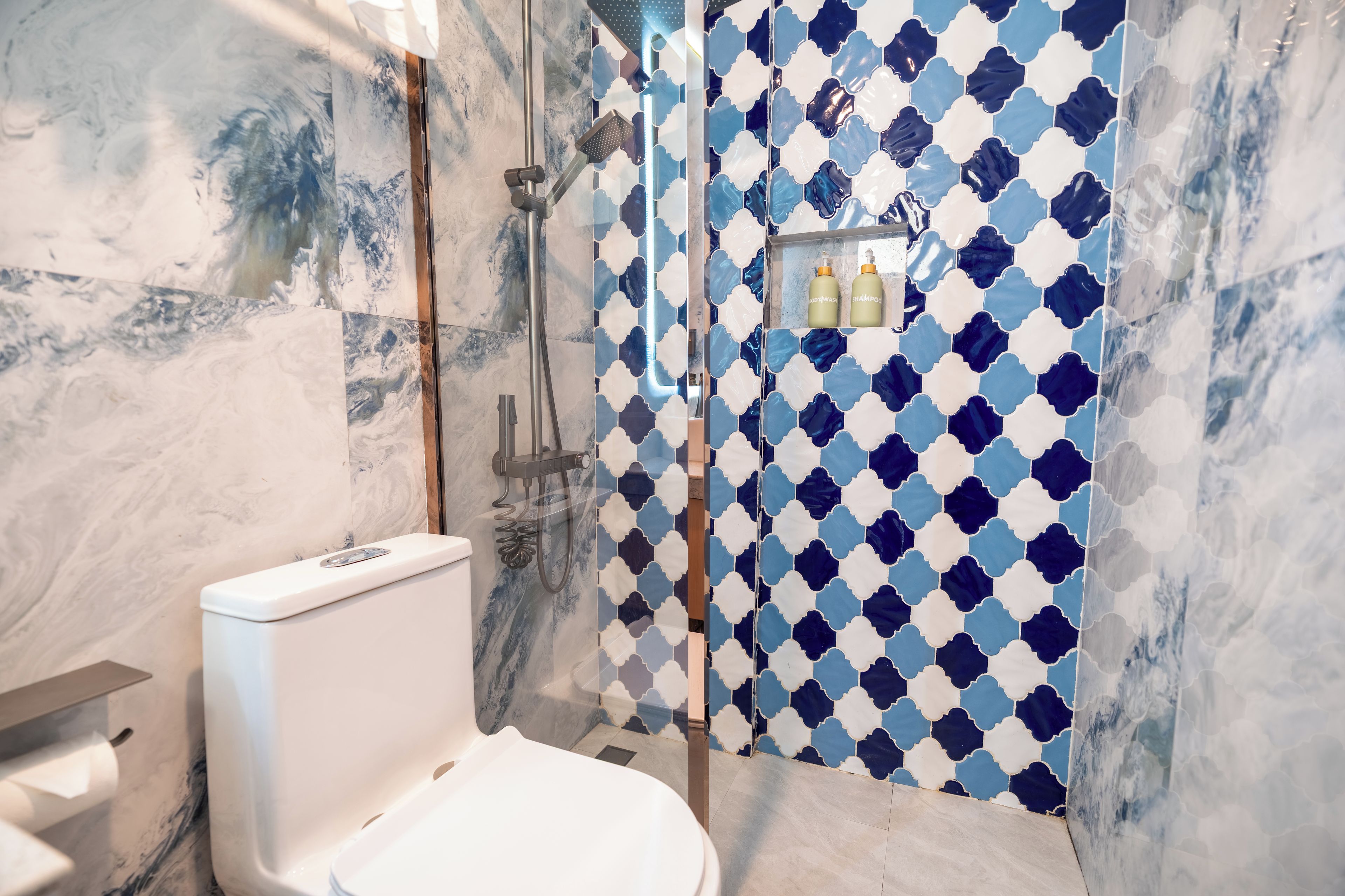 A bathroom with a marble floor, a toilet, a shower with a blue and white patterned wall, and a shelf with toiletries.