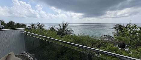 Un balcón con vista al océano, barandilla de cristal y una mesa con una silla.