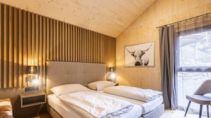 Een slaapkamer met een wand van houten latten, een bed met wit beddengoed, een nachtkastje met een lamp en een raam met uitzicht.