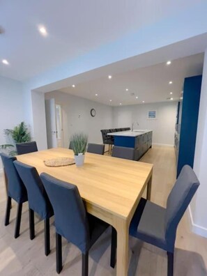 A modern dining area with a wooden table and six chairs, a small plant, and a kitchen in the background. - The Gatehouse & Lodge | Large Group Retreat (Holt)