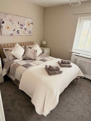 Un dormitorio con cama, almohadas, mesita de noche, lámpara, ventana con persianas y una obra de arte floral en la pared.