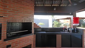 An outdoor kitchen area with a brick oven, a sink, and a window overlooking a residential area. - Recanto Reino Encantado 1 (Olímpia)