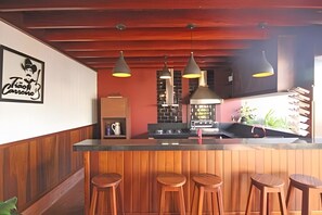 Zona de bar amb tamborets de fusta, il·luminació penjant i una cuina al fons.