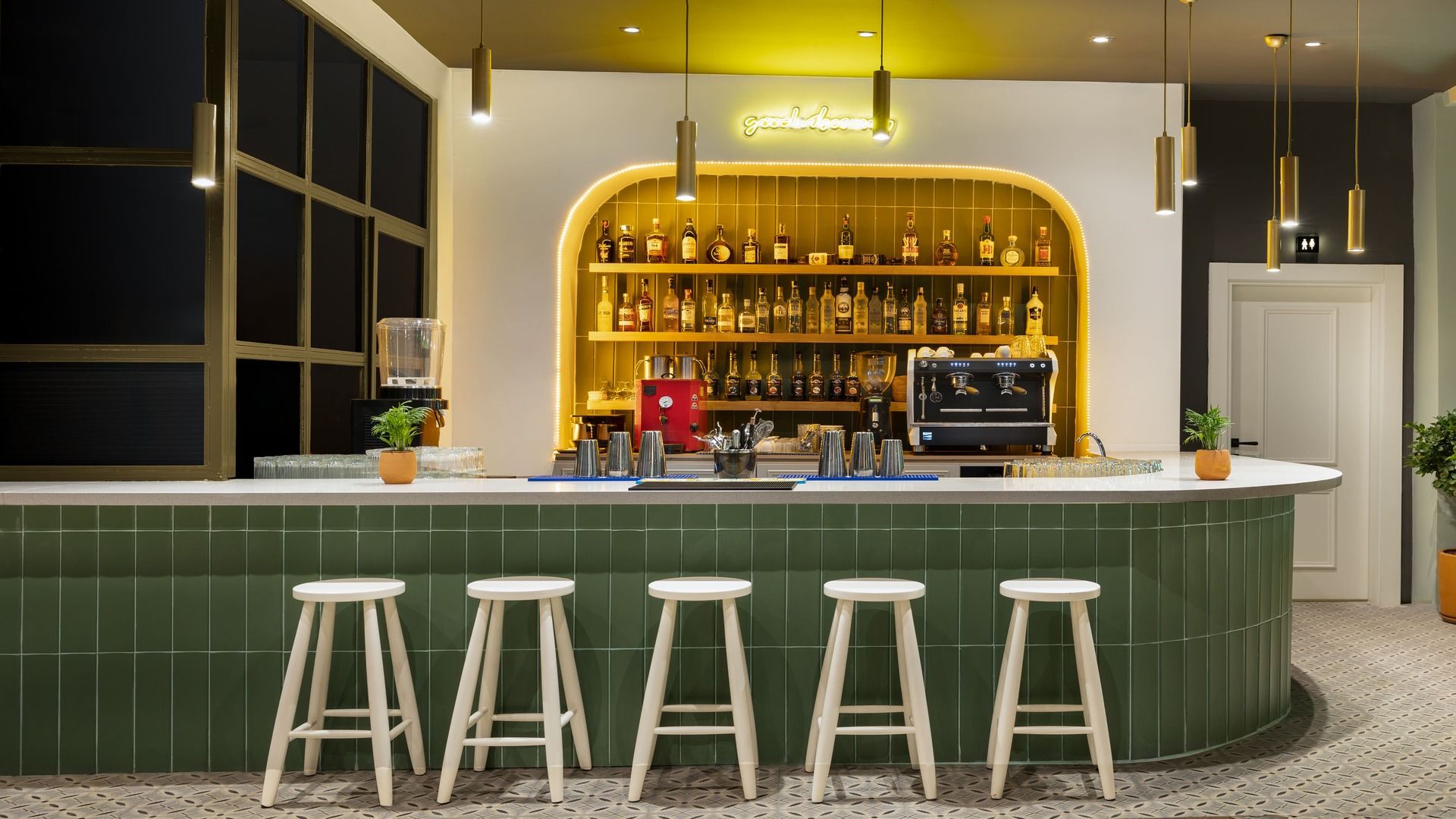 Una zona de bar con una barra verde, taburetes, una máquina de café y estantes llenos de botellas de licor.