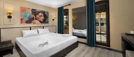 A hotel room with a large bed, a painting on the wall, a desk with a computer, and a view through a glass door.