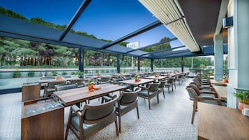 Un área de comedor al aire libre con varias mesas y sillas, un recinto de vidrio y vistas a zonas verdes.
