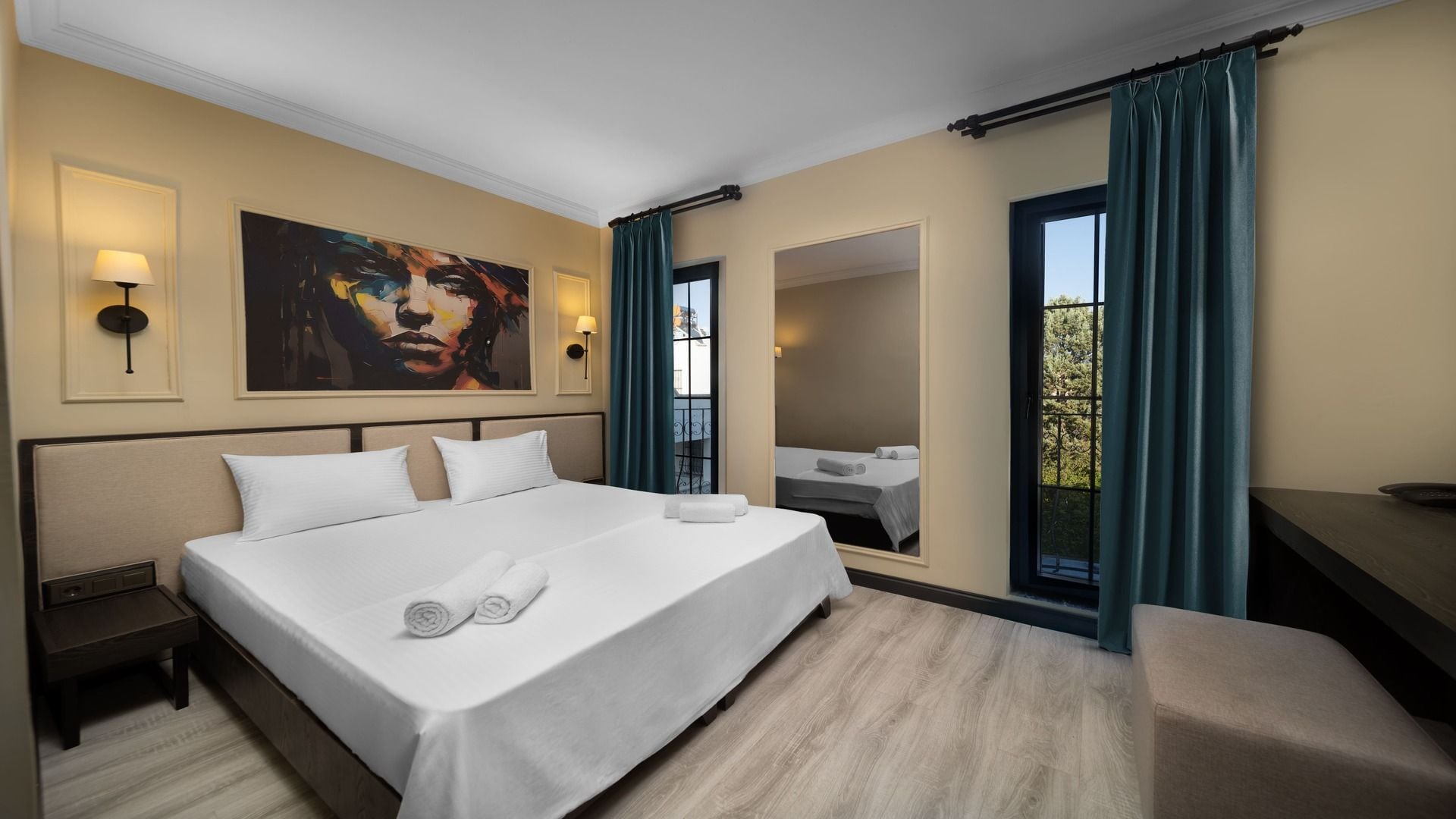 A hotel room with a large bed, a bedside table, a nightstand, a painting on the wall, a mirror, and a balcony with curtains.