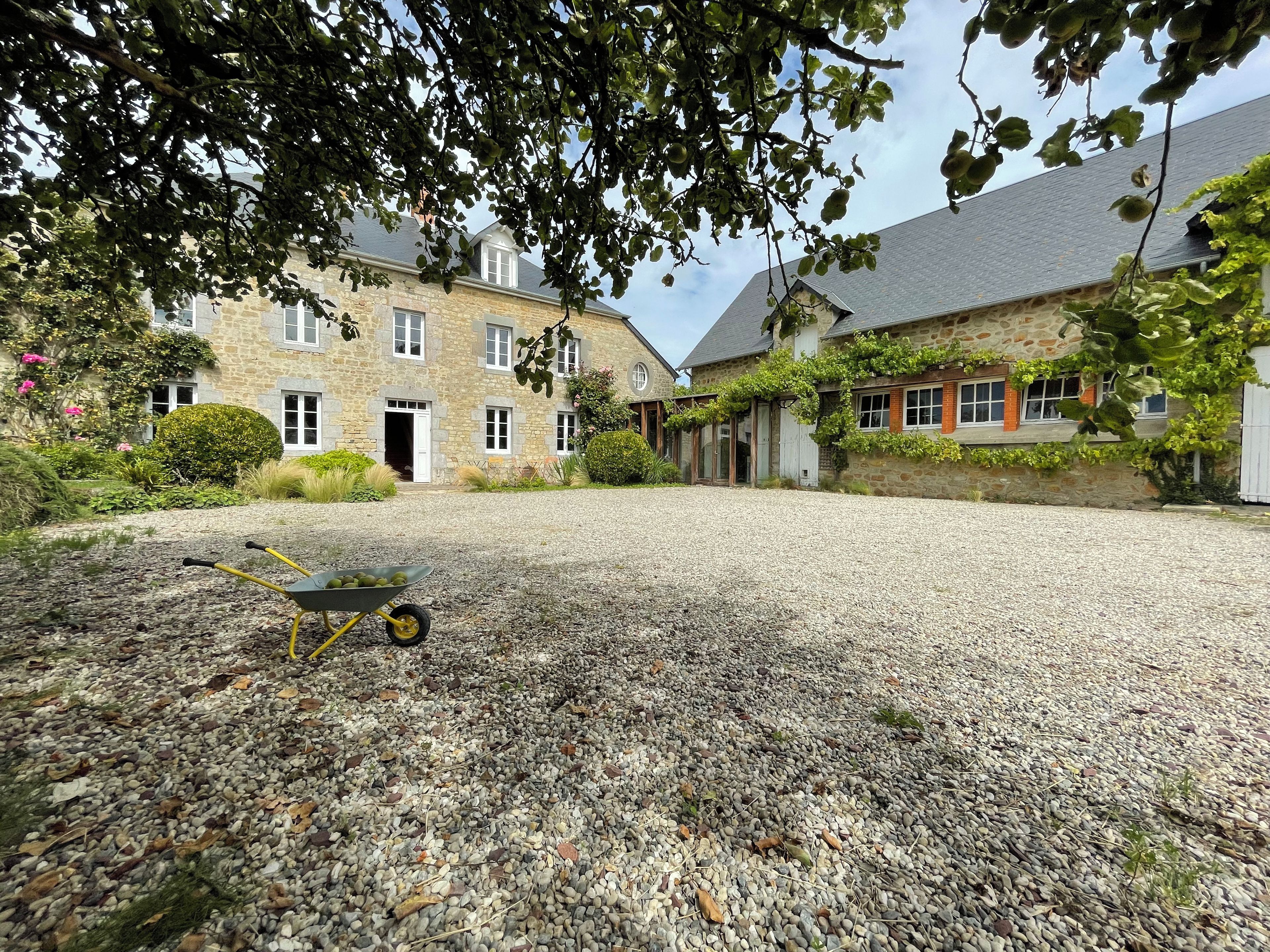 A courtyard with a gravel surface, a wheelbarrow, and stone buildings with windows and doors.