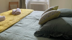 Una cama bien hecha con una manta amarilla y toallas enrolladas, dos almohadas estampadas y vistas a una ventana con cortinas.