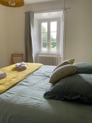 Una cama bien hecha con una manta amarilla y toallas enrolladas, dos almohadas estampadas y vistas a una ventana con cortinas.