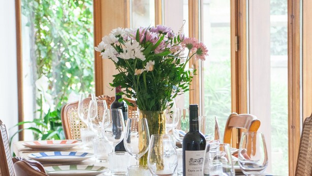 Una mesa de comedor dispuesta con platos, vasos y una botella de vino, rodeada de sillas de mimbre.
