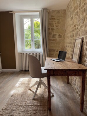 Une pièce avec une table en bois, un ordinateur portable, une chaise, une fenêtre avec des rideaux et un mur en pierre.
