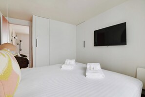 Quarto de hotel com uma cama, duas toalhas brancas, uma televisão na parede e um guarda-roupa.