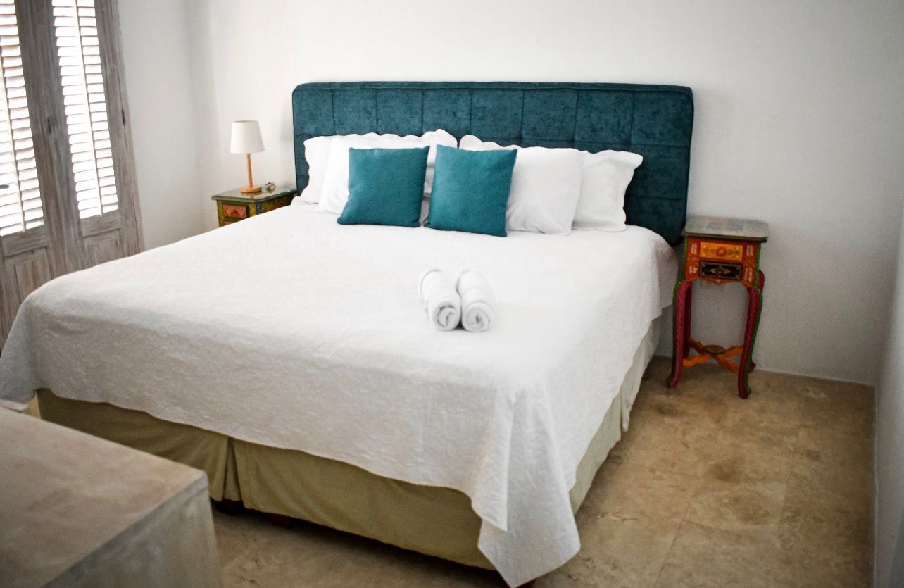 Una cama bien tendida con una colcha blanca y almohadas color turquesa, una mesita de noche con una lámpara y una pequeña mesa con una radio.