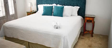 A neatly made bed with a white bedspread and teal pillows, a wooden nightstand with a lamp, and a small table with a radio.