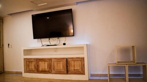 Ein Wohnzimmer mit einem flachbildfernseher an einer weißen Wand, Holzkredänzen darunter und zwei leeren Holzhochentischen.