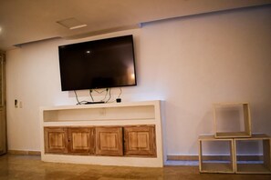 Een woonkamer met een flatscreen televisie aan een witte muur, houten kasten eronder en twee lege houten bijzettafels.