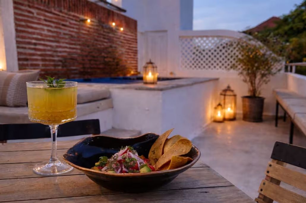 Een tafel met een drankje en een schaal chips en salsa, voor een achtergrond van een bakstenen muur en een terras.