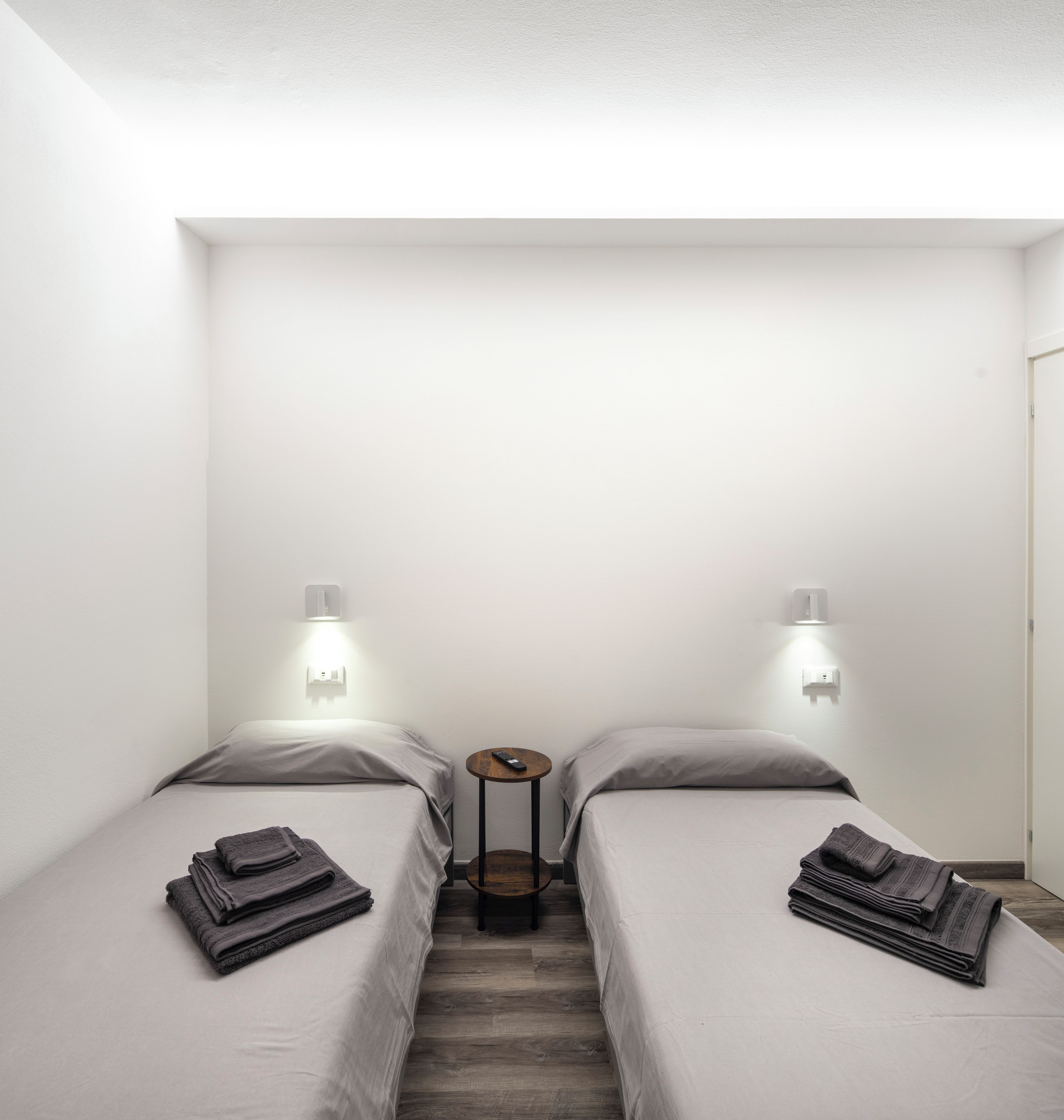Una habitación pequeña y moderna con dos camas, cada una con una almohada y una toalla doblada. Hay una mesita redonda pequeña entre las camas con un control remoto encima.