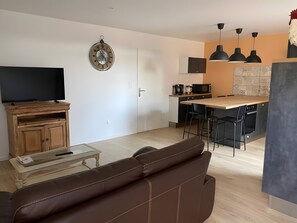 A modern living room with a brown leather sofa, a flat-screen TV on a wooden cabinet, and a dining area with a wooden table and chairs.