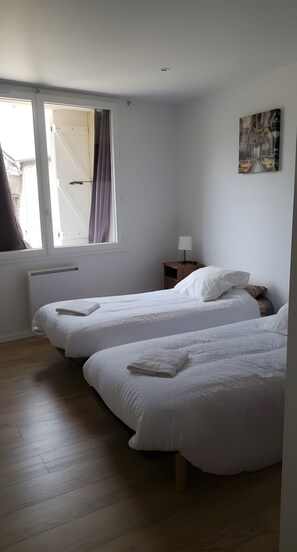 A room with two beds, a window with curtains, a radiator, a nightstand with a lamp, and a wall-mounted painting.