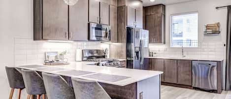 A modern kitchen with a central island, bar stools, stainless steel appliances, and pendant lighting.