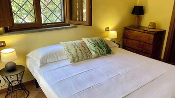 Una cama bien hecha con un cojín estampado, una lámpara de noche y una mesita de madera con una lámpara y un sombrero.