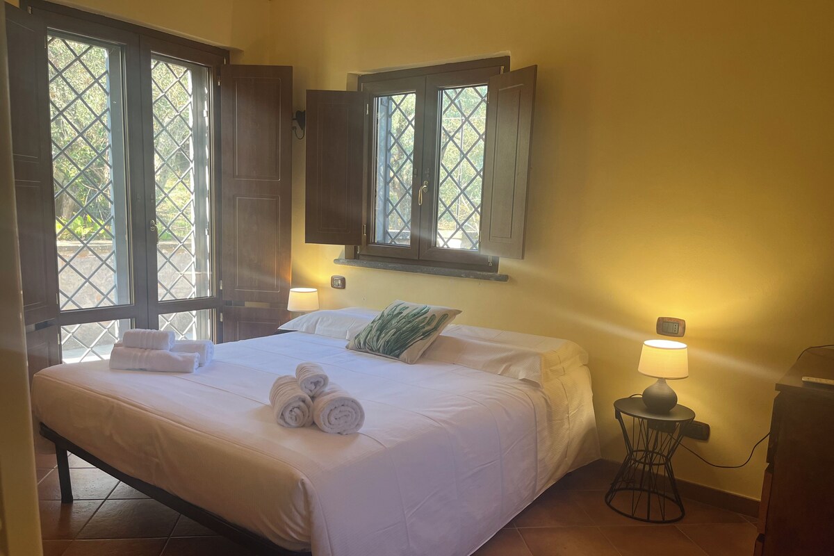 Habitación de hotel con cama, dos lámparas de noche, ventana con persianas y un reloj en la pared.