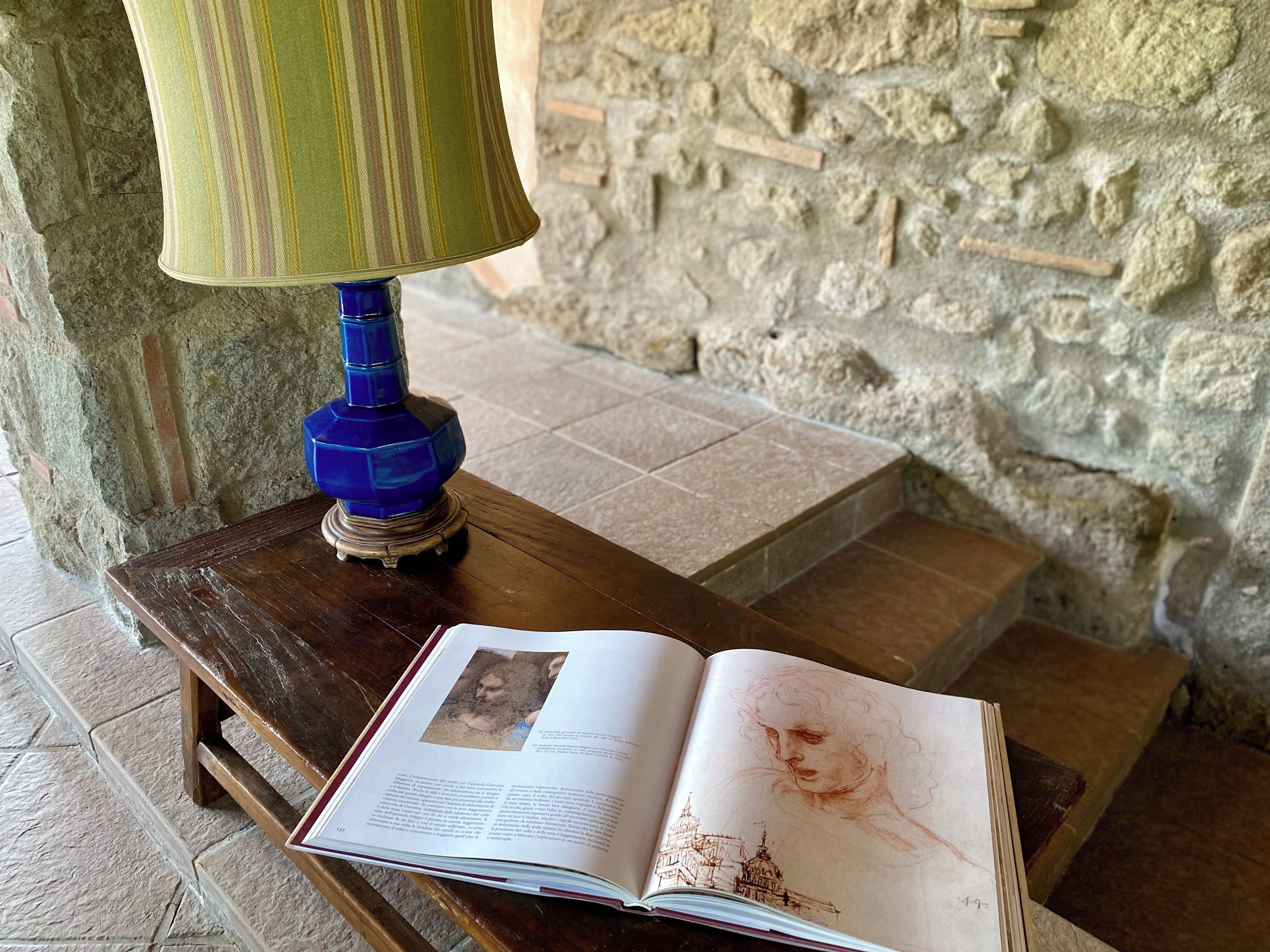 A book open on a wooden table with a blue lamp beside it, set against a stone wall.