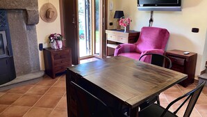 A living room with a stone fireplace, a wooden dining table, a pink armchair, a flat-screen TV mounted on the wall, and a small wooden cabinet. - Cà Fondi  (Lazio)